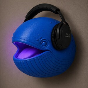 Op de afbeelding: Een blauwe walvisvormige luidspreker met een paars oplichtende mond en een zwarte koptelefoon. De walvis heeft een textuur en is bevestigd aan een neutraal gekleurde muur.