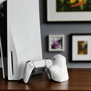 May include: A white PlayStation 5 console and controller are displayed on a wooden surface. A white ghost-shaped object is next to the controller. The background includes framed artwork.