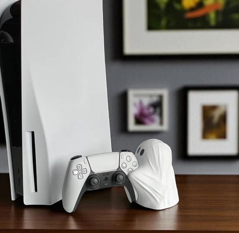 May include: A white PlayStation 5 console and controller are displayed on a wooden surface. A white ghost-shaped object is next to the controller. The background includes framed artwork.