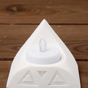 May include: A white, triangular candle holder with a decorative geometric design. It features a small, battery-operated LED candle. The holder is made of a solid, opaque material and is designed to hold a single tea light.