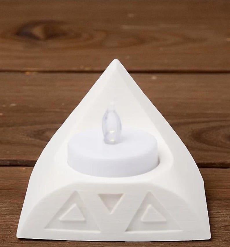 May include: A white, triangular candle holder with a decorative geometric design. It features a small, battery-operated LED candle. The holder is made of a solid, opaque material and is designed to hold a single tea light.