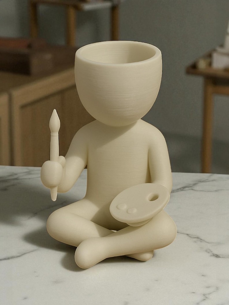 May include: A cream-colored figurine of a person seated cross-legged, holding a paintbrush and a palette. The figure has a large, open head, suggesting a vase or planter. The overall design is minimalist and artistic.