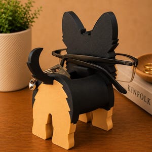 Pode incluir: Um porta-&oacute;culos em forma de c&atilde;o preto e castanho com cauda e porta-chaves. O c&atilde;o est&aacute; segurando um par de &oacute;culos. O suporte &eacute; feito de madeira e fica em uma superf&iacute;cie de madeira. Uma pequena planta em um vaso est&aacute; ao fundo.
