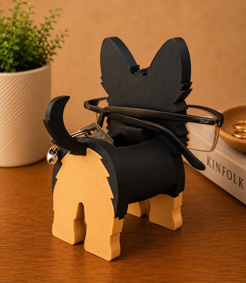 Pode incluir: Um porta-&oacute;culos em forma de c&atilde;o preto e castanho com cauda e porta-chaves. O c&atilde;o est&aacute; segurando um par de &oacute;culos. O suporte &eacute; feito de madeira e fica em uma superf&iacute;cie de madeira. Uma pequena planta em um vaso est&aacute; ao fundo.