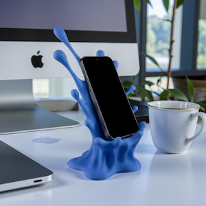 May include: A blue phone stand in a splash design holds a black smartphone. The stand is on a white desk, with a laptop and a white mug with a gold rim in the background.