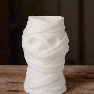 May include: A white, mummy-themed candle holder. The candle holder is shaped like a skull wrapped in bandages. The bandages are textured and create a layered effect. The candle holder is a decorative item.