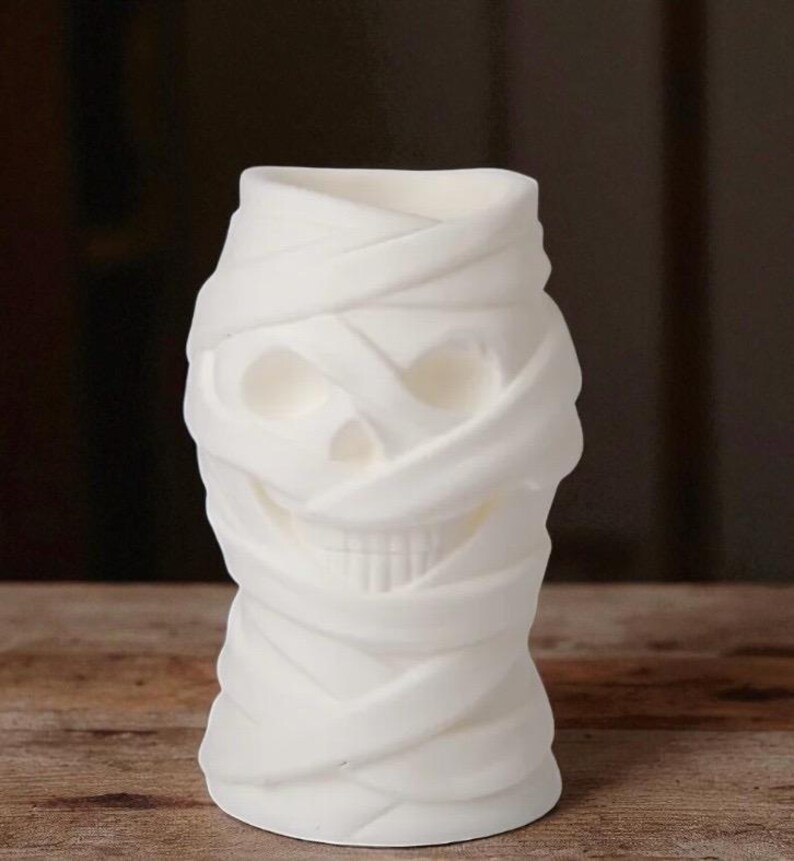 May include: A white, mummy-themed candle holder. The candle holder is shaped like a skull wrapped in bandages. The bandages are textured and create a layered effect. The candle holder is a decorative item.