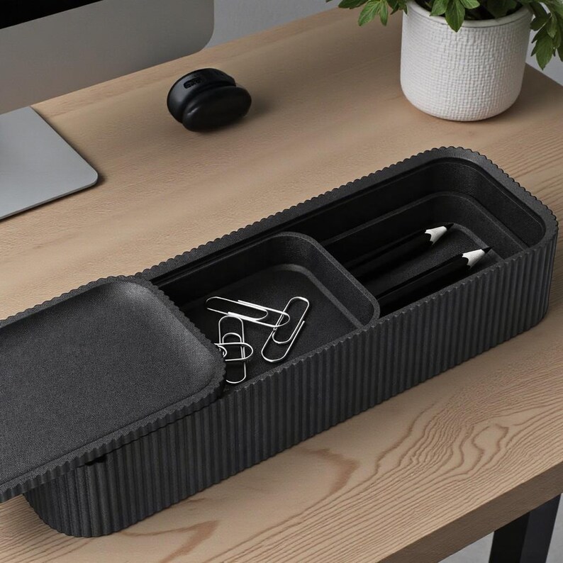 May include: A black desk organizer with a textured, ribbed design. The organizer contains a small tray with paper clips and a compartment with two pencils. The organizer sits on a light wood desk.