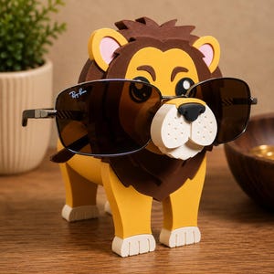 May include: A whimsical lion-shaped eyeglass holder in yellow, brown, and white, with a pair of sunglasses resting on its nose. The sunglasses have dark lenses and a silver frame. The lion has a friendly expression and is made of a foam-like material.