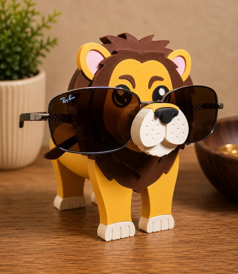 May include: A whimsical lion-shaped eyeglass holder in yellow, brown, and white, with a pair of sunglasses resting on its nose. The sunglasses have dark lenses and a silver frame. The lion has a friendly expression and is made of a foam-like material.