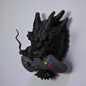 May include: A black dragon sculpture with a video game controller. The dragon's head is detailed with textured scales and horns. The controller has colored buttons and directional controls. The sculpture is mounted on a white wall.