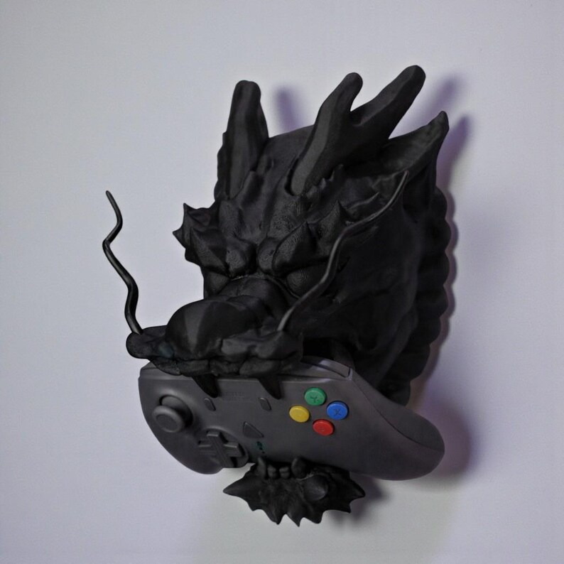 May include: A black dragon sculpture with a video game controller. The dragon's head is detailed with textured scales and horns. The controller has colored buttons and directional controls. The sculpture is mounted on a white wall.