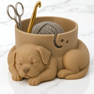 Puede incluir: Un cuenco de hilo beige con un dise&ntilde;o de cachorro durmiendo. El cuenco contiene una madeja de lana gris, un ganchillo dorado y unas tijeras plateadas. El cuenco tiene una abertura curva para el hilo.