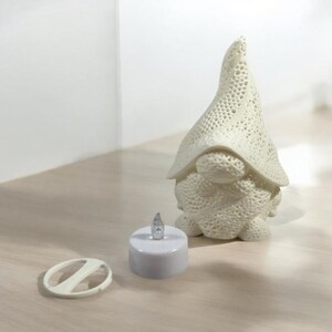 May include: A cream-colored gnome figurine with a textured, openwork hat and body. Beside it is a small, white, battery-operated candle with a flickering LED. A white remote control is also present.
