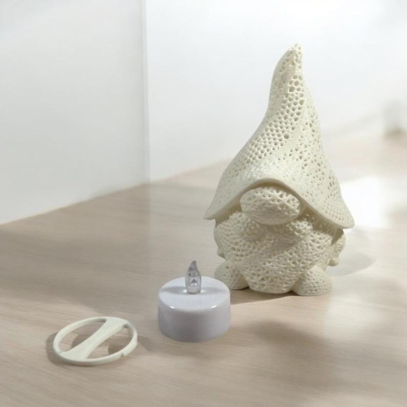 May include: A cream-colored gnome figurine with a textured, openwork hat and body. Beside it is a small, white, battery-operated candle with a flickering LED. A white remote control is also present.