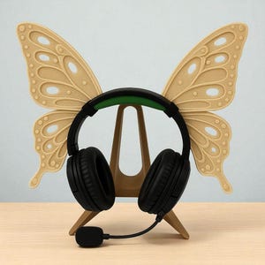 Puede incluir: Un auricular negro con un acento verde descansa sobre un soporte de madera con forma de mariposa. Las alas de la mariposa son beige con patrones detallados. El soporte está sobre una superficie de madera clara.