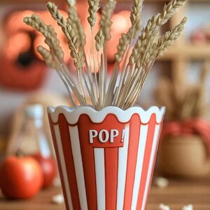 May include: A popcorn-themed container with red and white stripes, holding dried wheat stalks. The container has a scalloped rim and the word "POP!" in white. The arrangement sits on a wooden surface.