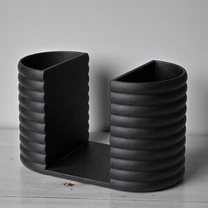 May include: A matte black bookend with a unique design. The bookend features two curved sides with a ridged texture, connected by a flat base. The sides have a semi-circular shape, and the overall design is modern and minimalist.