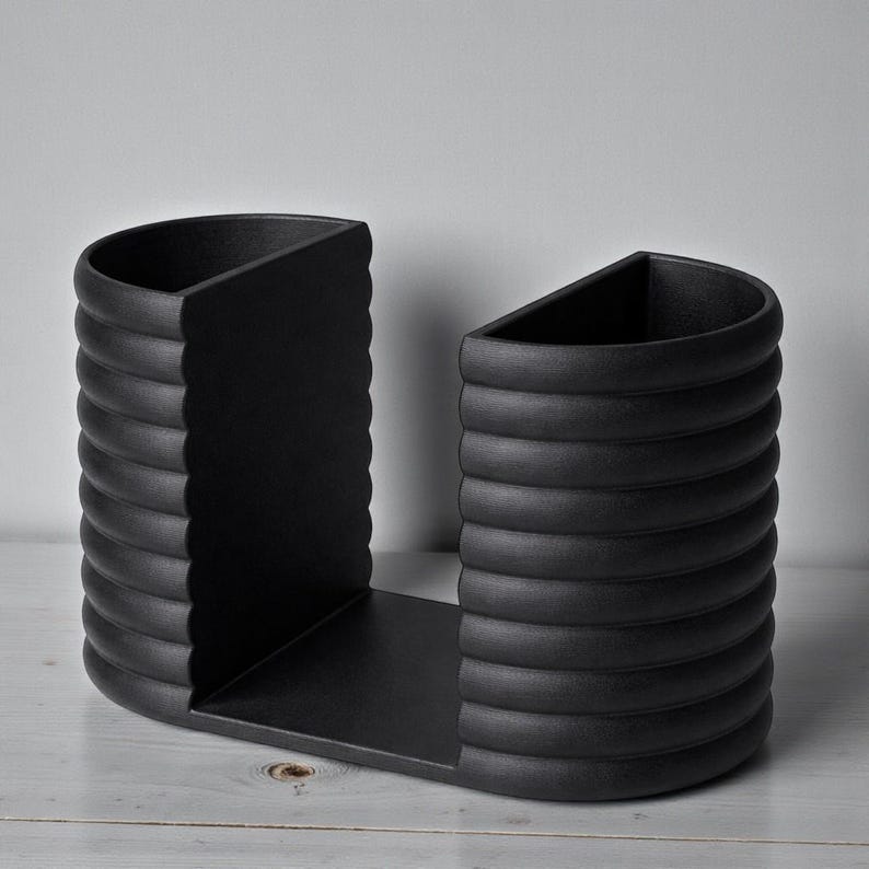 May include: A matte black bookend with a unique design. The bookend features two curved sides with a ridged texture, connected by a flat base. The sides have a semi-circular shape, and the overall design is modern and minimalist.