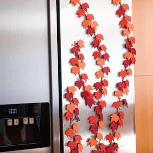 May include: Decorative fall leaf garland in shades of red and orange, arranged in vertical strands. The garland is attached to a stainless steel refrigerator door, creating a seasonal autumn display. The leaves are paper or felt.