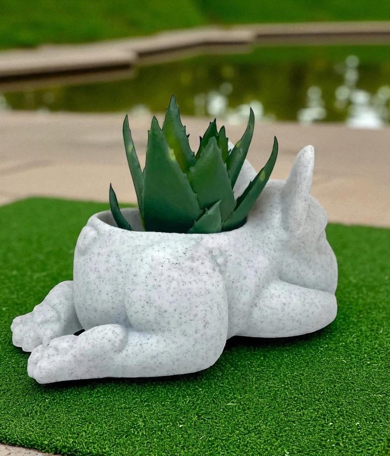 May include: A speckled white ceramic planter shaped like a reclining bulldog, with a green succulent plant inside. The planter has a textured finish and is set on a green surface.