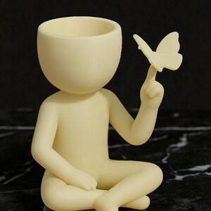 Puede incluir: Figura de color crema de una persona sentada, con la cabeza hueca y una mariposa posada en un dedo. La figura es lisa y tiene un dise&ntilde;o minimalista, adecuada para la decoraci&oacute;n del hogar o como un regalo &uacute;nico.