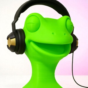 May include: A vibrant green frog ornament wearing black and gold headphones. The frog has its eyes closed and a large smile. The headphones have gold detailing. The sculpture is set against a white and pink backdrop.