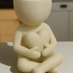 May include: A cream-colored ceramic planter shaped like a person holding a heart. The figure is seated with legs crossed and arms embracing the heart. A small plant sprouts from the head of the planter. The design is minimalist and modern.