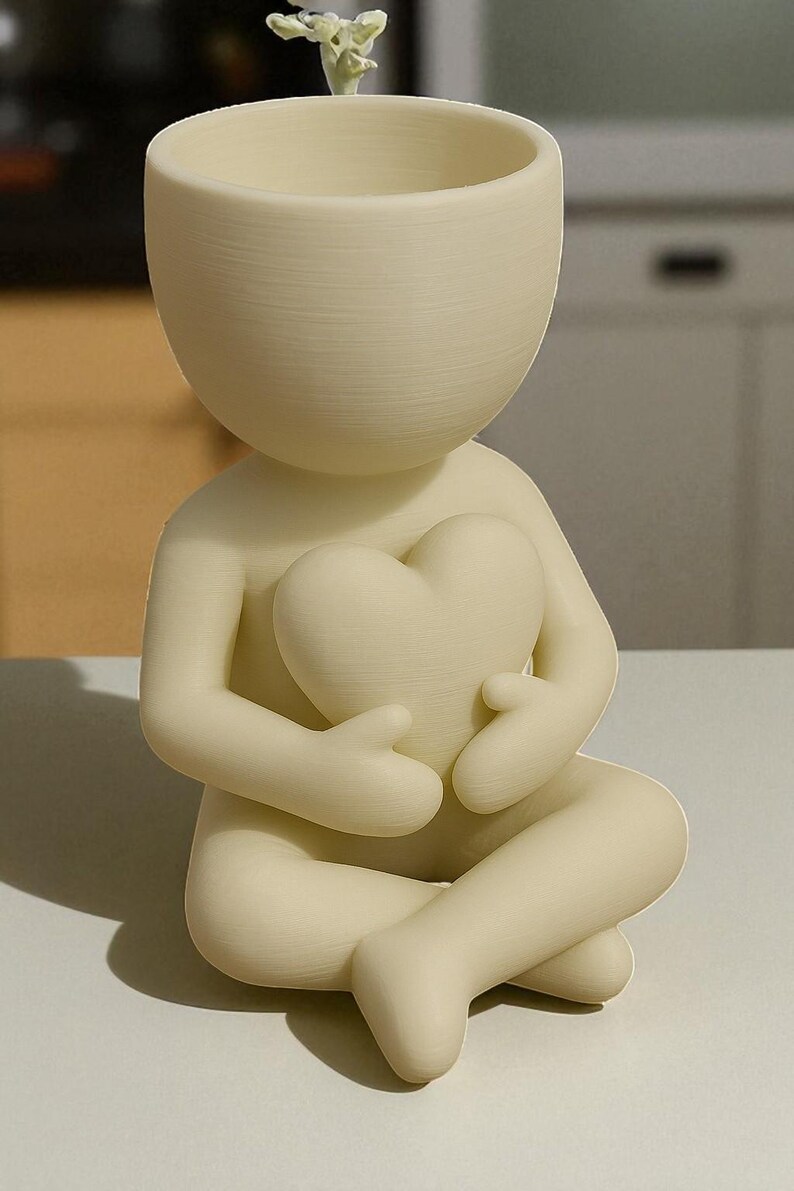 May include: A cream-colored ceramic planter shaped like a person holding a heart. The figure is seated with legs crossed and arms embracing the heart. A small plant sprouts from the head of the planter. The design is minimalist and modern.
