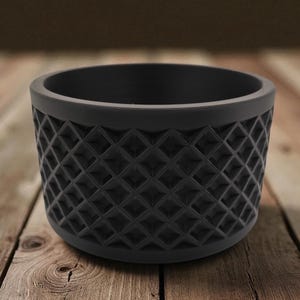 May include: A black, cylindrical planter with a geometric diamond pattern around the exterior. The planter sits on a wooden surface. The planter is approximately 4 inches in diameter and 3 inches tall.