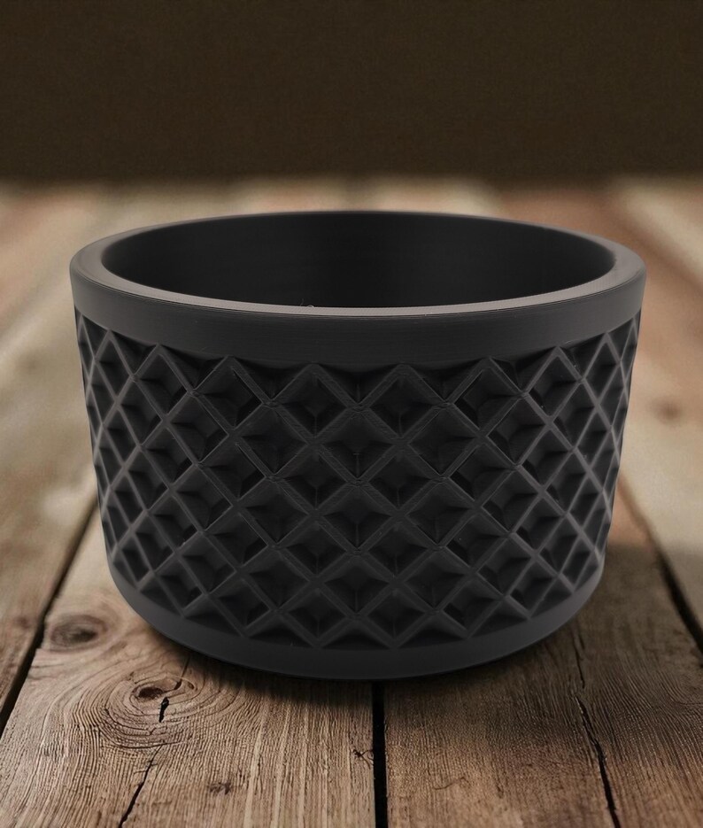 May include: A black, cylindrical planter with a geometric diamond pattern around the exterior. The planter sits on a wooden surface. The planter is approximately 4 inches in diameter and 3 inches tall.