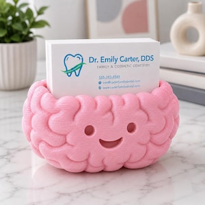 May include: A pink, brain-shaped business card holder with a smiling face. The holder contains a stack of white business cards with the text "Dr. Emily Carter, DDS, Family & Cosmetic Dentistry". The holder is on a light-colored surface.