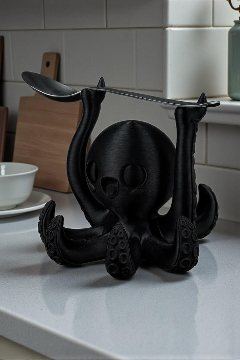 Puede incluir: Un soporte negro con forma de pulpo para utensilios de cocina. El pulpo tiene una cabeza redonda, ojos grandes y tent&aacute;culos. Una cuchara plateada descansa sobre los brazos del pulpo. El soporte es de material negro mate.