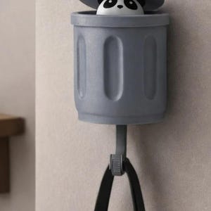 May include: A gray panda bear trash can-shaped wall hook. The trash can has a lid that is slightly open, revealing the panda's face. The hook is attached to a black strap.