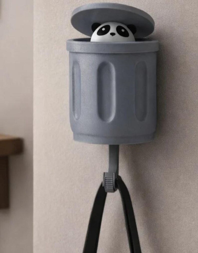 May include: A gray panda bear trash can-shaped wall hook. The trash can has a lid that is slightly open, revealing the panda's face. The hook is attached to a black strap.