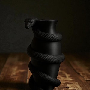 以下が含まれることがあります： 黒色のつや消し花瓶。蛇が巻き付くデザインで、蛇の頭が縁にあります。花瓶は暗い背景に置かれ、木製の表面にあります。