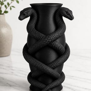May include: Black vase with two snakes coiled around it. The snakes' bodies are textured, and their heads are positioned at the top of the vase. The vase has a wide opening and a round base. The background is a light marble surface.