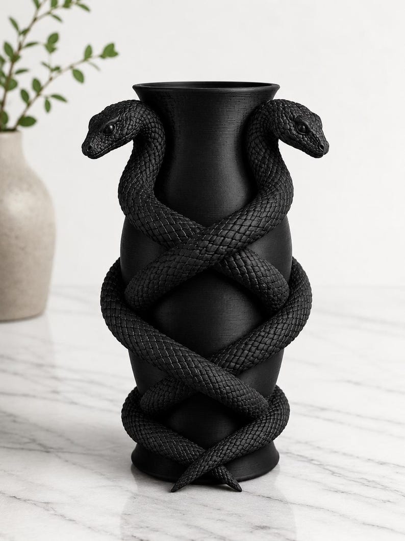 May include: Black vase with two snakes coiled around it. The snakes' bodies are textured, and their heads are positioned at the top of the vase. The vase has a wide opening and a round base. The background is a light marble surface.