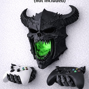 May include: A black, horned skull-shaped wall mount with a green glowing interior, flanked by two game controller holders. The text "mounts to wall with screws (not included)" is above the skull.