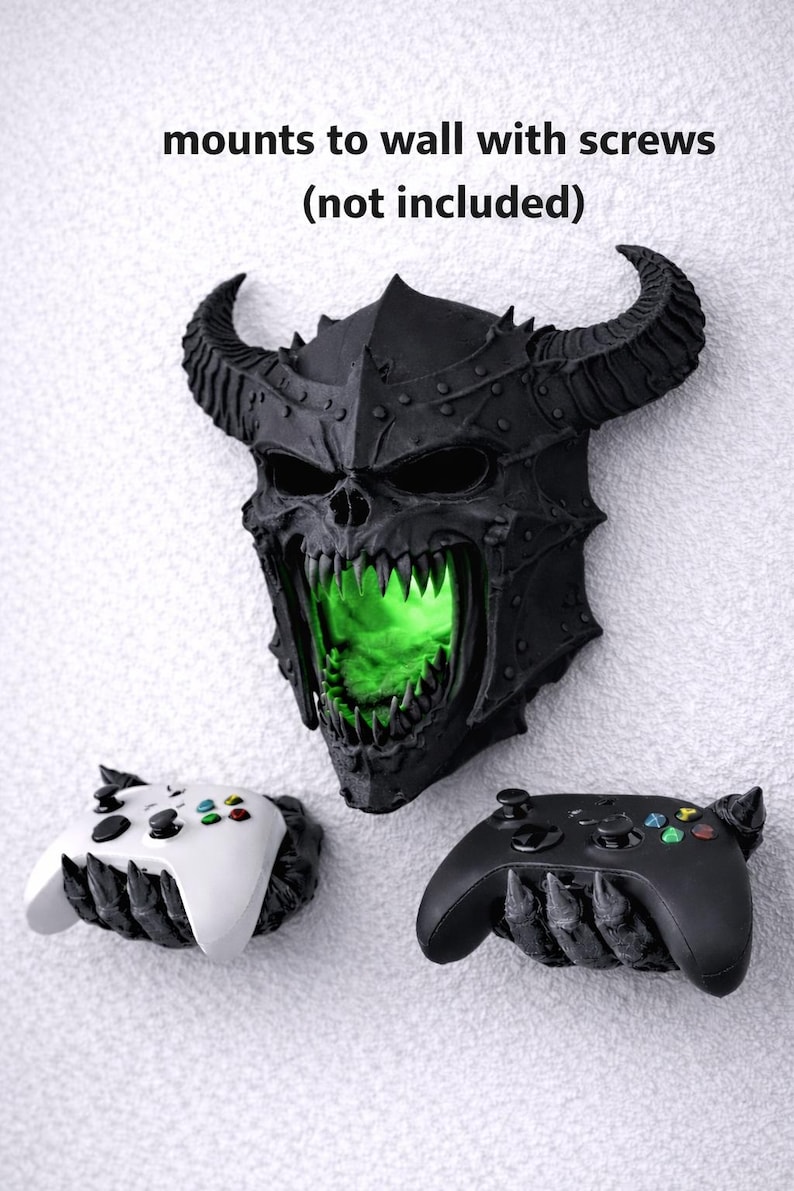 May include: A black, horned skull-shaped wall mount with a green glowing interior, flanked by two game controller holders. The text "mounts to wall with screws (not included)" is above the skull.