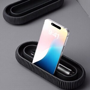 May include: A black, oval-shaped desk organizer with a ribbed exterior. A smartphone displaying 9:41 is placed in the organizer, along with two pens. Another organizer is visible in the background. The items are on a gray surface.