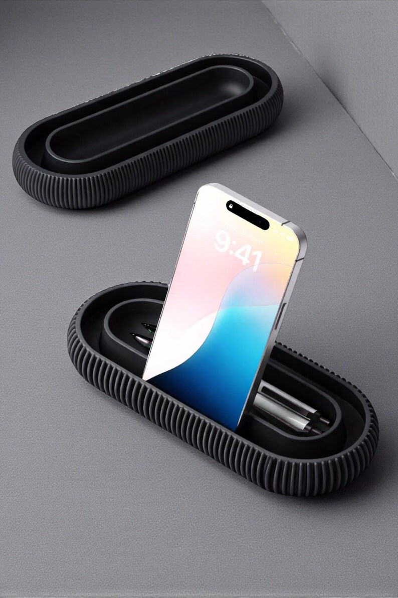 May include: A black, oval-shaped desk organizer with a ribbed exterior. A smartphone displaying 9:41 is placed in the organizer, along with two pens. Another organizer is visible in the background. The items are on a gray surface.