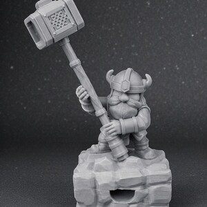 May include: A grey figurine of a dwarf wielding a large hammer. The dwarf is wearing a horned helmet and stands on a rocky base. The hammer head features a lattice design. The figurine is made of a solid, matte material.
