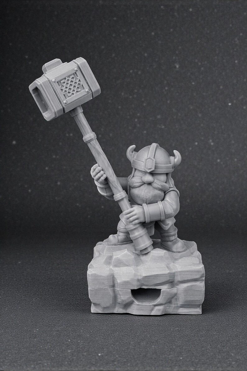 May include: A grey figurine of a dwarf wielding a large hammer. The dwarf is wearing a horned helmet and stands on a rocky base. The hammer head features a lattice design. The figurine is made of a solid, matte material.