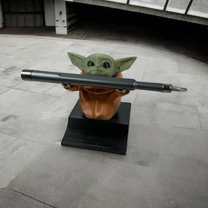 Può includere: Una statuetta verde e beige di Baby Yoda che tiene un cacciavite grigio scuro. La statuetta indossa una veste arancione ed è montata su una base nera. Il cacciavite è lungo circa 25 cm.