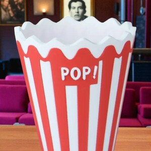 May include: A red and white striped popcorn container with a scalloped rim. The word "POP!" is printed in white on the front. The container is designed to resemble a classic popcorn bucket.