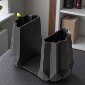 May include: A dark gray, geometric desk organizer with two angled compartments. The organizer has a textured surface and a modern, angular design. A yellow pen is visible in one compartment. The organizer sits on a dark gray surface.