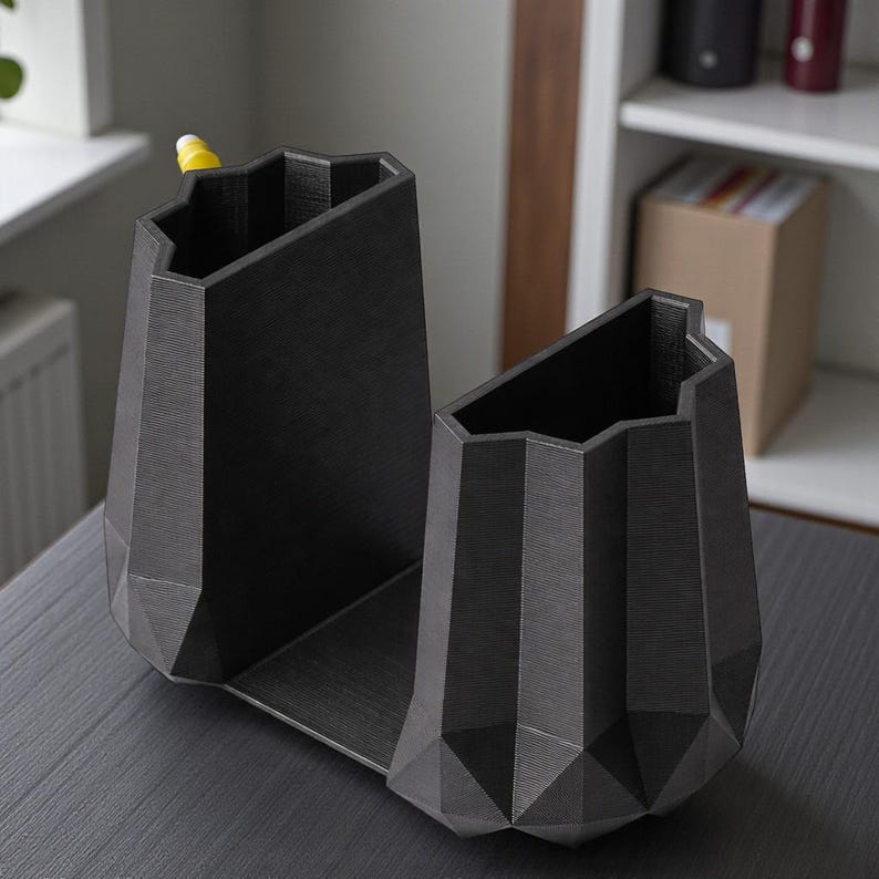 May include: A dark gray, geometric desk organizer with two angled compartments. The organizer has a textured surface and a modern, angular design. A yellow pen is visible in one compartment. The organizer sits on a dark gray surface.