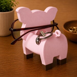 May include: A pink pig-shaped eyeglasses and key holder. The holder has a light pink body with brown feet and a curly tail. Eyeglasses are resting on the pig's head, and keys are in the keyhole. A decorative item for a desk or nightstand.