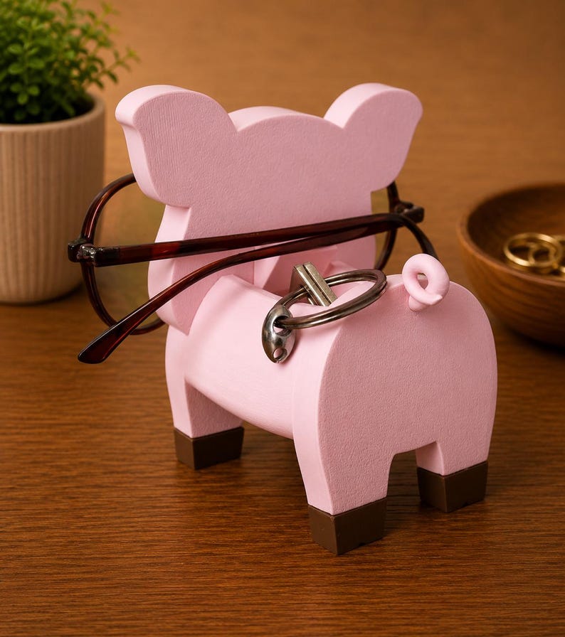 May include: A pink pig-shaped eyeglasses and key holder. The holder has a light pink body with brown feet and a curly tail. Eyeglasses are resting on the pig's head, and keys are in the keyhole. A decorative item for a desk or nightstand.